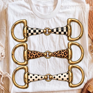 Op de afbeelding: Een wit t-shirt met een grafische print van drie horizontale paardenbitten in goud. Het bovenste bit heeft een zwart-wit dambordpatroon, het middelste bit heeft een bruin-zwarte luipaardprint en het onderste bit heeft zwarte stippen.