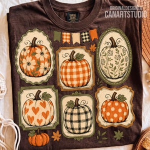 May include: A dark brown t-shirt featuring a graphic print of six pumpkins in various patterns, including floral, gingham, plaid, and polka dots, framed in decorative borders. Above the pumpkins, a row of small colorful pennant flags is displayed.