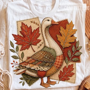 Peut inclure: Un t-shirt blanc avec une impression graphique représentant une oie entourée de feuilles d'automne. Le motif utilise un style patchwork avec des textures rappelant la toile de jute, le tartan et le tissu cousu dans des tons orange, rouge, marron et vert.