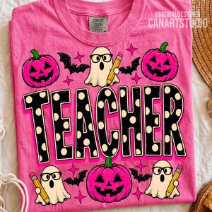 May include: A bright pink t-shirt featuring a graphic print with the word TEACHER in large black letters with white polka dots. The design includes three white ghosts wearing black glasses, three pink jack-o'-lanterns, bats, and yellow pencils.