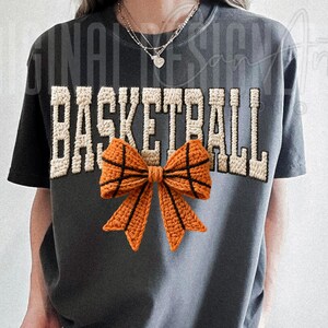 Op de afbeelding: Donkergrijs T-shirt met het woord "BASKETBALL" in een cr&egrave;mekleurig, getextureerd lettertype. Een gehaakte oranje strik, ontworpen om op een basketbal te lijken, is gecentreerd onder de tekst. Het shirt heeft korte mouwen en een ronde hals.