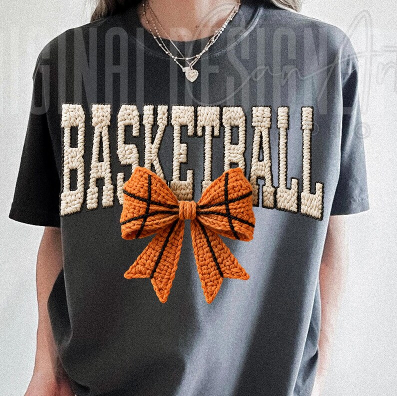 Op de afbeelding: Donkergrijs T-shirt met het woord "BASKETBALL" in een cr&egrave;mekleurig, getextureerd lettertype. Een gehaakte oranje strik, ontworpen om op een basketbal te lijken, is gecentreerd onder de tekst. Het shirt heeft korte mouwen en een ronde hals.