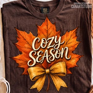 May include: A dark brown t-shirt featuring a large, detailed orange and yellow maple leaf graphic. Centered on the leaf is the white text "COZY SEASON" in a cursive script font. A golden-yellow ribbon bow is illustrated at the base of the leaf.