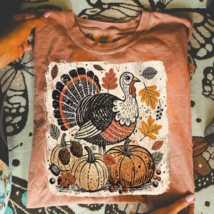 Pode incluir: Uma t-shirt cor de p&ecirc;ssego com um gr&aacute;fico tem&aacute;tico de A&ccedil;&atilde;o de Gra&ccedil;as. O design inclui um peru detalhado com cauda em leque, ab&oacute;boras e folhas de outono em tons de castanho, laranja e bege. A t-shirt est&aacute; dobrada, mostrando o gr&aacute;fico de forma proeminente.
