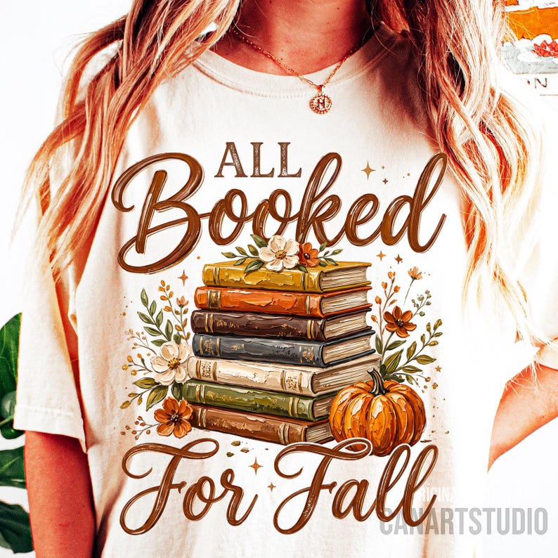 May include: Cream-colored t-shirt with the text "ALL Booked For Fall" in brown script. The design features a stack of books, flowers, and a pumpkin in autumnal colors. A perfect tee for book lovers.