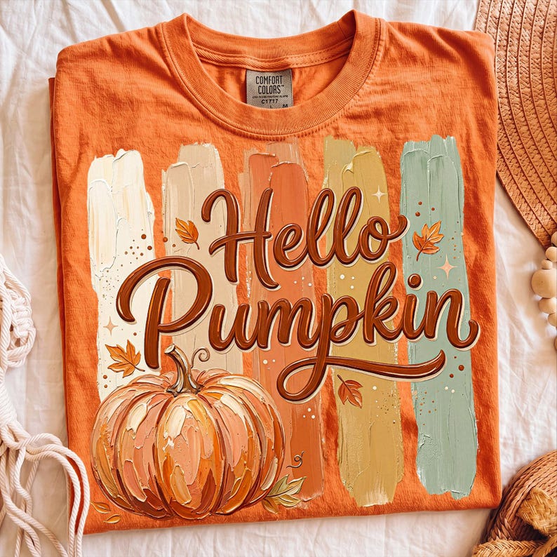 Puede incluir: Camiseta naranja con la frase "Hello Pumpkin" en una fuente decorativa, acompa&ntilde;ada de una ilustraci&oacute;n de calabaza y pinceladas coloridas. La camiseta es de la marca Comfort Colors.