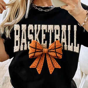 Op de afbeelding: Zwart T-shirt met het woord "BASKETBALL" in cr&egrave;mekleurige, getextureerde letters. Een oranje en zwarte gehaakte strik, die op een basketbal lijkt, is onder de tekst gecentreerd. Het shirt is een casual, sportief ontwerp.