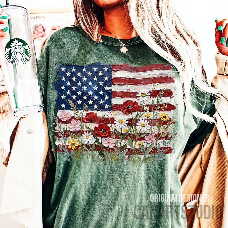 May include: Green crew neck t-shirt featuring a painted American flag design with red, white, and blue colors. The flag design includes white stars and red and white stripes, with colorful flowers at the bottom. The text "ORIGINAL DESIGNER CANVAS STUDIO" is visible.
