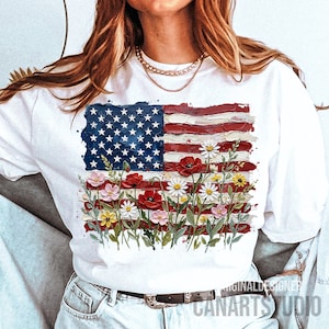 May include: White t-shirt featuring a painted American flag design with flowers. The flag's blue field has white stars, and the red stripes are accented with poppies, daisies, and other colorful blooms. The text "CANARTS STUDIO" is visible.