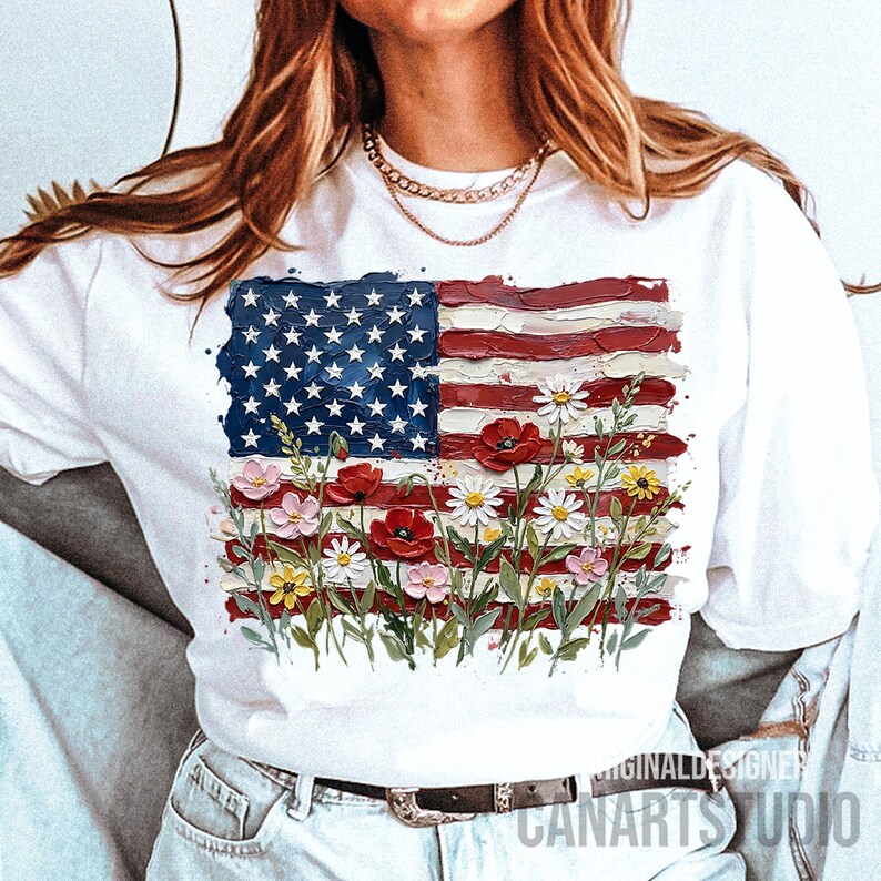 May include: White t-shirt featuring a painted American flag design with flowers. The flag's blue field has white stars, and the red stripes are accented with poppies, daisies, and other colorful blooms. The text "CANARTS STUDIO" is visible.