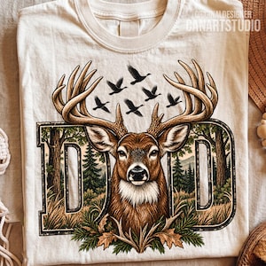 May include: A cream-colored t-shirt featuring a graphic of a deer head with large antlers centered between the letters D and D. The design includes a forest landscape with pine trees, mountains, and a flock of birds flying above the deer.