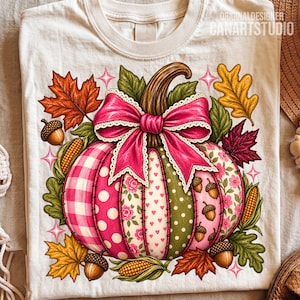 May include: A cream-coloured t-shirt featuring a central graphic of a pumpkin with patchwork-style panels in pink gingham, polka dots, and floral patterns. The pumpkin is topped with a large pink bow and surrounded by autumn leaves, acorns, and corn.