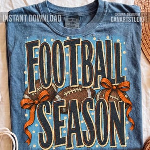 K&ouml;nnte beinhalten: Ein blaues T-Shirt mit dem Text FOOTBALL SEASON in dunkelblauen Buchstaben. Das Design zeigt braune Football-Grafiken, orangefarbene Schleifen und wei&szlig;e Punkte auf hellblauem Grund. Das T-Shirt hat einen Rundhalsausschnitt und ein Comfort Colors Etikett.