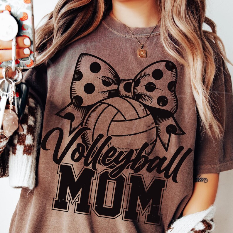 May include: Brown t-shirt with a black and white polka dot bow and a volleyball. The text "Volleyball Mom" is printed in black on the shirt.