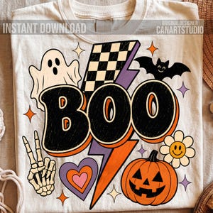 May include: A cream-colored t-shirt featuring a graphic print with the text BOO in large black letters. The design includes a white ghost, a black bat, a jack-o'-lantern, a skeleton hand making a peace sign, a checkered lightning bolt, and a flower.