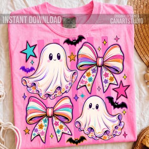 May include: A bright pink t-shirt featuring a graphic print of two white ghosts, two large bows with colorful floral and striped patterns, several small black bats, and various stars in shades of teal, orange, and pink against a striped background.