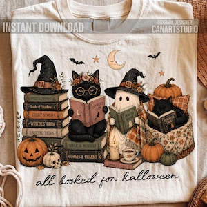 May include: A cream-colored t-shirt featuring a graphic of two black cats and a ghost wearing witch hats, reading books. The design includes pumpkins, a skull, a crescent moon, and the text "all booked for halloween" at the bottom.