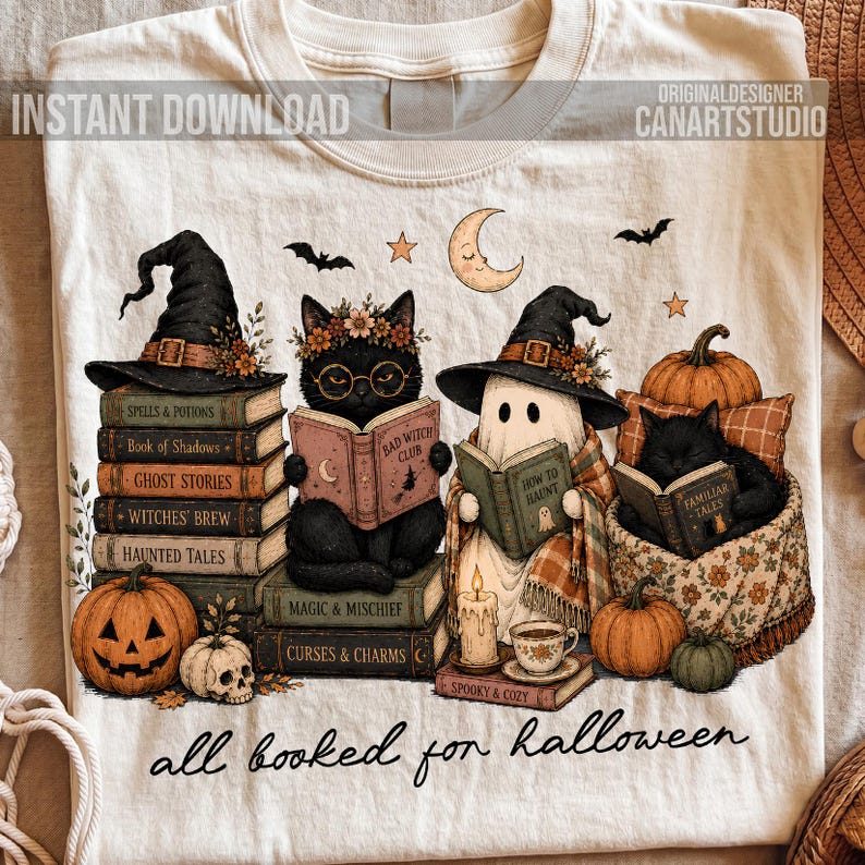May include: A cream-colored t-shirt featuring a graphic of two black cats and a ghost wearing witch hats, reading books. The design includes pumpkins, a skull, a crescent moon, and the text "all booked for halloween" at the bottom.