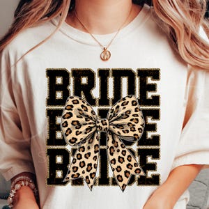 May include: White t-shirt with the text "BRIDE" in black with a gold glitter outline. A large leopard print bow is centred on the shirt.