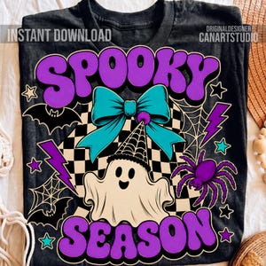 May include: A black t-shirt featuring a graphic print with the text "SPOOKY SEASON" in purple. The design includes a white ghost wearing a party hat, a large teal bow, a black and white checkered background, bats, spiderwebs, and a purple spider.