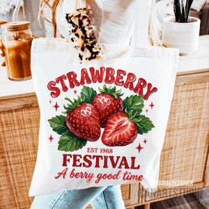 May include: A white tote bag with a colourful strawberry design. The design features the word "STRAWBERRY" in red, three strawberries, green leaves, and the words "EST 1968" and "FESTIVAL A berry good time".