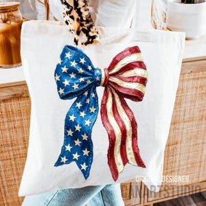 May include: A white tote bag with a patriotic bow design in red, white, and blue, reminiscent of the American flag. The bow features blue sections with gold stars and red and white stripes. The words "ORIGINAL DESIGNER CANARTSTUDIO" are printed on the bottom right.