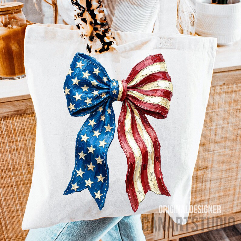 May include: A white tote bag with a patriotic bow design in red, white, and blue, reminiscent of the American flag. The bow features blue sections with gold stars and red and white stripes. The words "ORIGINAL DESIGNER CANARTSTUDIO" are printed on the bottom right.