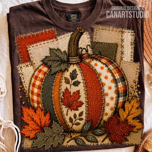 May include: A dark brown t-shirt featuring a large graphic of a pumpkin composed of various textured fabric patches in orange, cream, and green. The pumpkin is surrounded by detailed autumn leaves and set against a background of stitched fabric squares.