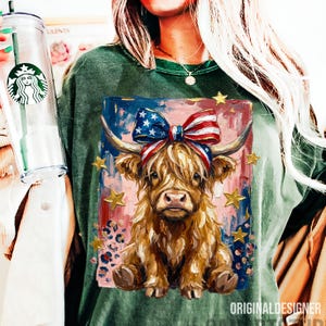 Peut inclure: T-shirt vert &agrave; manches longues avec une illustration de vache des Highlands orn&eacute;e d'un n&oelig;ud aux couleurs du drapeau am&eacute;ricain et d'&eacute;toiles dor&eacute;es. L'&oelig;uvre est plac&eacute;e sur un fond rouge, blanc et bleu. Le t-shirt porte le texte "ORIGINALDESIGNER".
