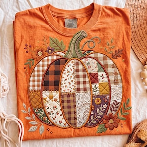 Pode incluir: Camiseta laranja Comfort Colors com um design de ab&oacute;bora bordada. A ab&oacute;bora &eacute; feita de v&aacute;rios padr&otilde;es de tecido, incluindo xadrez e estampas florais. A camisa tem gola redonda e mangas curtas.