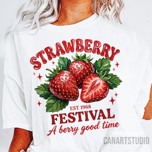 May include: White t-shirt with a strawberry-themed graphic. The design showcases three red strawberries, green leaves, and the words "STRAWBERRY FESTIVAL" with "EST 1968" and "A berry good time" in red text. The design is by CANARTSTUDIO.