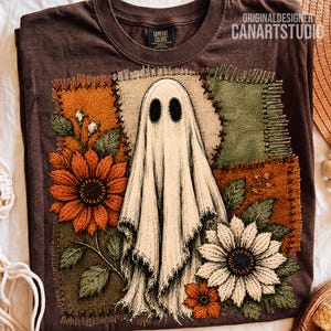 Op de afbeelding: Een donkerbruin t-shirt met een centrale afbeelding van een witte geest omringd door borduurstijl patches in oranje, groen en bruin. Het ontwerp bevat gedetailleerde zonnebloemen en bladeren, met de tekst ORIGINALDESIGNER CANARTSTUDIO.