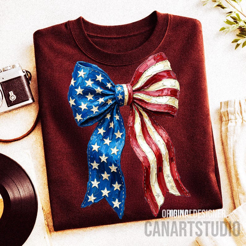 May include: A maroon crew neck sweatshirt with a large bow design. The bow features a patriotic design with blue sections with white stars and red and white stripes. The text "ORIGINAL DESIGNER CANARTSTUDIO" is printed at the bottom of the design.