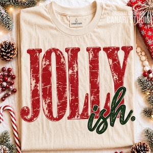 Può includere: Una t-shirt color crema con la parola JOLLY in grandi lettere maiuscole rosse dall'aspetto usurato e la parola ish in un carattere corsivo verde scuro più piccolo. La maglietta presenta un'etichetta sul collo con il testo CANARTSTUDIO.