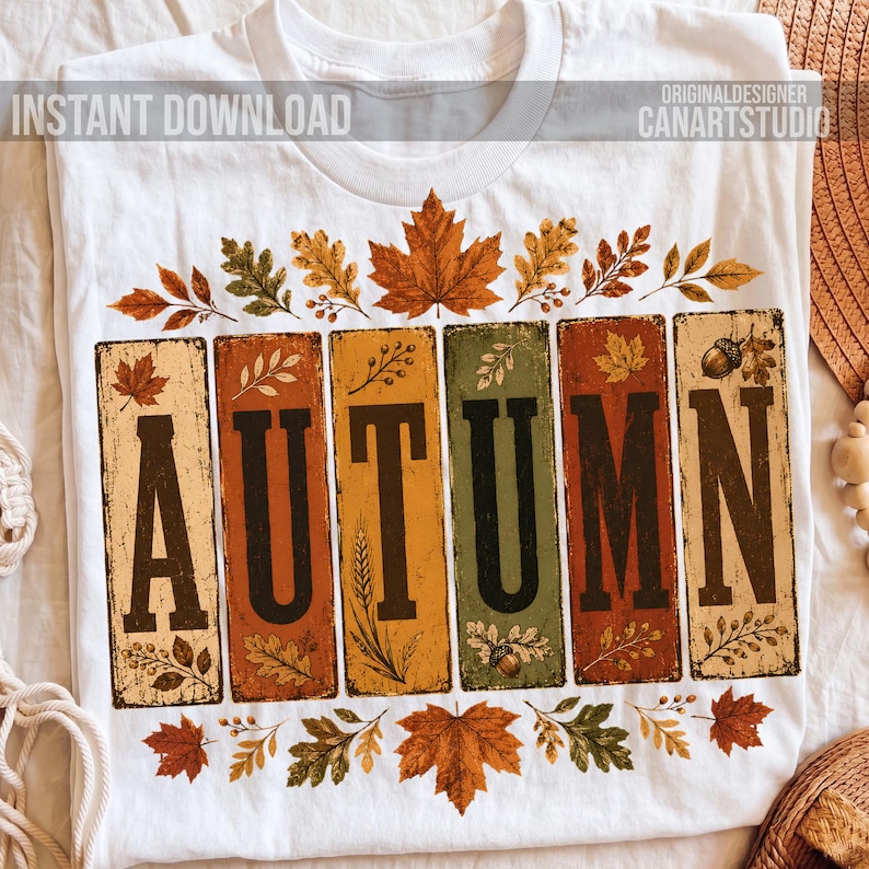 May include: A white t-shirt featuring the word AUTUMN in large black letters, with each letter set inside a vertical rectangular panel of varying colors including orange, yellow, and green, decorated with illustrations of fall leaves and acorns.