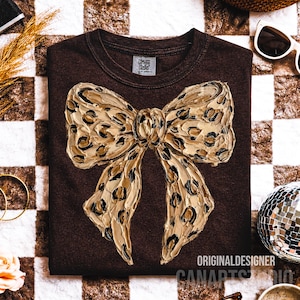 May include: A brown sweatshirt featuring a large, textured bow with a leopard print design. The bow is in shades of gold and black. The text "ORIGINAL DESIGNER CANARTS STUDIO" is printed at the bottom of the sweatshirt.