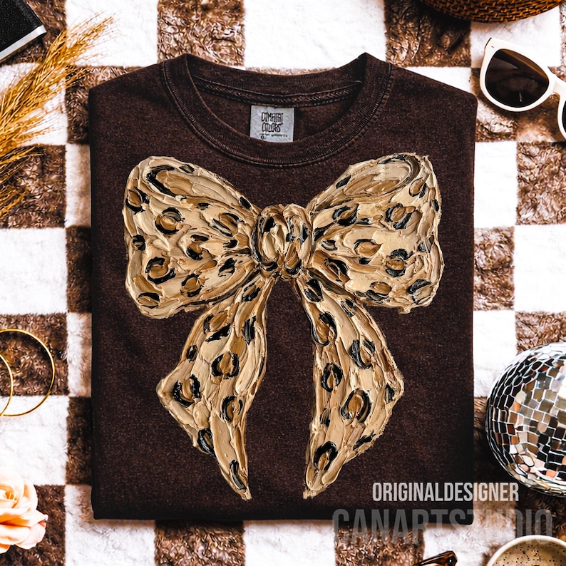 May include: A brown sweatshirt featuring a large, textured bow with a leopard print design. The bow is in shades of gold and black. The text "ORIGINAL DESIGNER CANARTS STUDIO" is printed at the bottom of the sweatshirt.