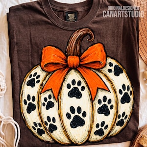 May include: A dark brown t-shirt featuring a large graphic of a cream-colored pumpkin with black paw prints and a bright orange bow on top. The shirt has a Comfort Colors label at the neck and the text ORIGINALDESIGNER CANARTSTUDIO.