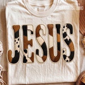 May include: A cream-colored t-shirt featuring the word JESUS in large, block letters. Each letter is filled with a patchwork of brown, black, and white cow-print patterns and is outlined with a border of small, white, round beads.
