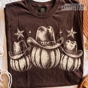 Può includere: Una t-shirt a girocollo marrone scuro con una stampa grafica di tre zucche, ognuna che indossa un cappello da cowboy. Il design include due stelle e due stelle a quattro punte. Il testo ORIGINALDESIGNER CANARTSTUDIO è stampato in alto a destra.