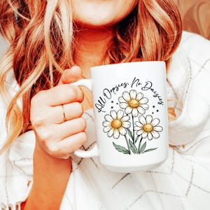 May include: White ceramic mug featuring a daisy floral design with the text "All Oopsies, No Daisies." The mug has a large handle and is filled with a beverage. The daisies have yellow centers and white petals.