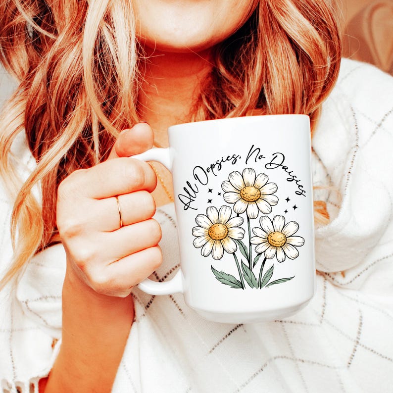 May include: White ceramic mug featuring a daisy floral design with the text "All Oopsies, No Daisies." The mug has a large handle and is filled with a beverage. The daisies have yellow centers and white petals.