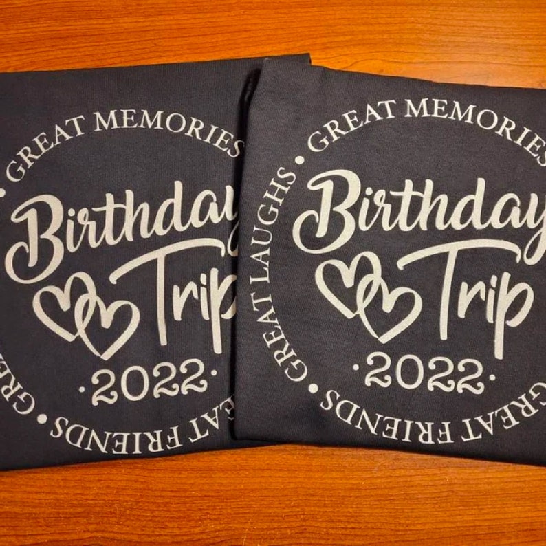 May include: Two black t-shirts with white text that reads "Great Memories. Birthday Trip. Great Laughs. 2022. Great Friends."  The text is in a circle with two hearts in the center.