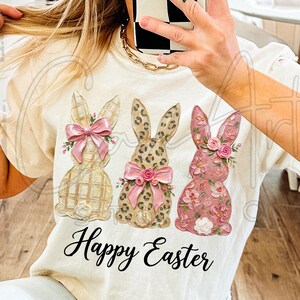 Puede incluir: Camiseta color crema con tres conejos decorativos de Pascua. Los conejos tienen diferentes patrones y colores, incluyendo cuadros, estampado de leopardo y dise&ntilde;os florales. Cada conejo tiene un lazo rosa y detalles florales. Las palabras "Happy Easter" est&aacute;n escritas en cursiva.