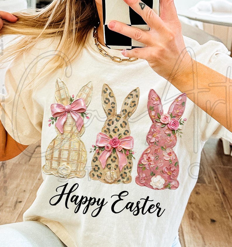Puede incluir: Camiseta color crema con tres conejos decorativos de Pascua. Los conejos tienen diferentes patrones y colores, incluyendo cuadros, estampado de leopardo y dise&ntilde;os florales. Cada conejo tiene un lazo rosa y detalles florales. Las palabras "Happy Easter" est&aacute;n escritas en cursiva.
