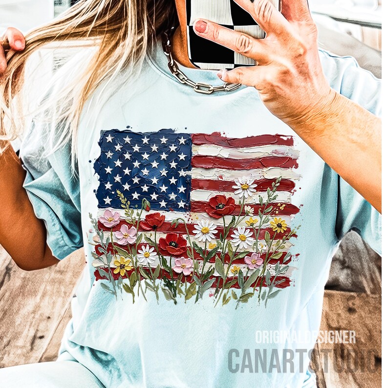 May include: Light blue t-shirt featuring a painted American flag design with red and white stripes, a blue star field, and colorful flowers. The shirt has short sleeves and a crew neck. The text "ORIGINAL DESIGNER CANARTSTUDIO" is visible.