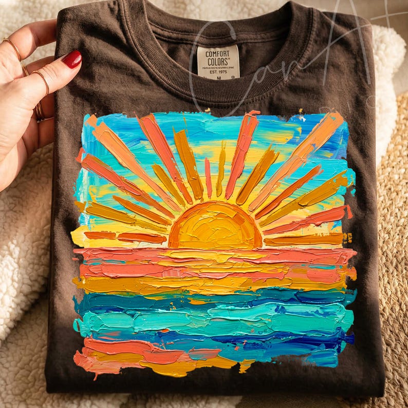 May include: Dark brown t-shirt featuring a vibrant sunset design. The artwork depicts a sun with orange and yellow rays over a colorful ocean, with shades of blue, orange, and yellow. The shirt has a Comfort Colors tag.