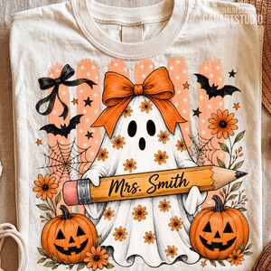 Puede incluir: Una camiseta color crema con el gr&aacute;fico de un fantasma con un lazo naranja grande, sosteniendo un l&aacute;piz amarillo con el texto Mrs. Smith. El dise&ntilde;o incluye calabazas naranjas, murci&eacute;lagos negros, telara&ntilde;as y flores peque&ntilde;as sobre un fondo a rayas.