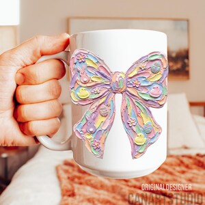 May include: White ceramic mug featuring a large, colorful bow design with Easter-themed embellishments. The bow is decorated with pastel eggs, bunnies, and flowers. The mug has a handle and the text "ORIGINALDESIGNER CANARTSTUDIO" is visible.