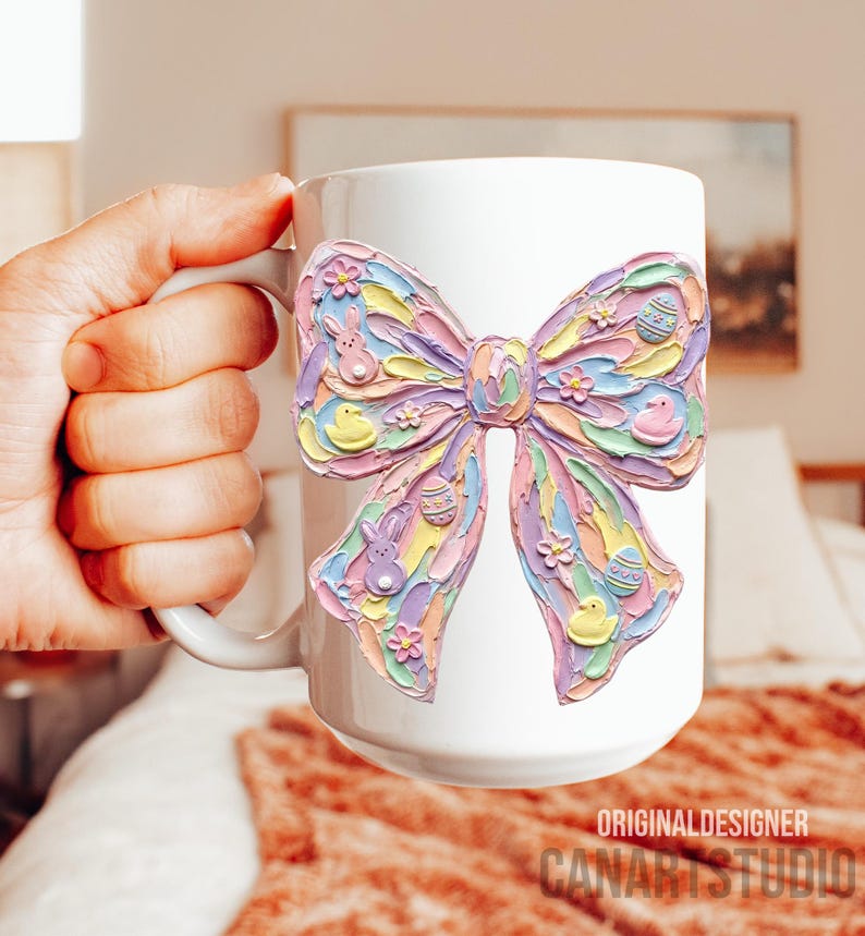 May include: White ceramic mug featuring a large, colorful bow design with Easter-themed embellishments. The bow is decorated with pastel eggs, bunnies, and flowers. The mug has a handle and the text "ORIGINALDESIGNER CANARTSTUDIO" is visible.
